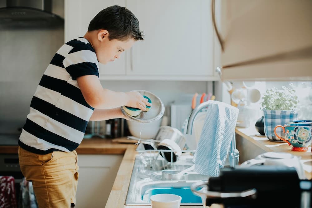 Young man with Down syndrome focused on washing a dish at a kitchen sink, with a running faucet and kitchen items around.